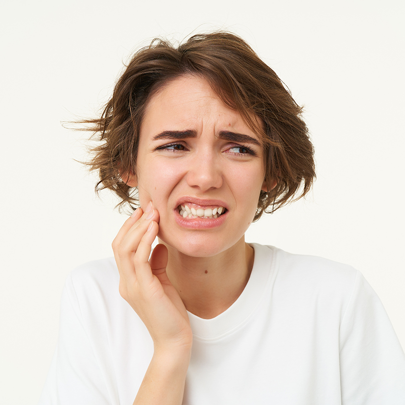 Close up of woman has a toothache, touches her teeth and frowns from painful discomfort, stands over white background Young woman experiencing discomfort from teeth grinding, highlighting cosmetic wellness challenges.